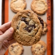holding a jacques torres chocolate chip cookie over pan of cookies.