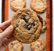 holding a jacques torres chocolate chip cookie over pan of cookies.