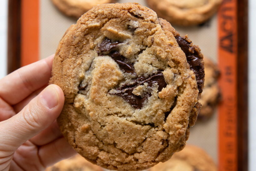 holding a jacques torres chocolate chip cookie over pan of cookies.
