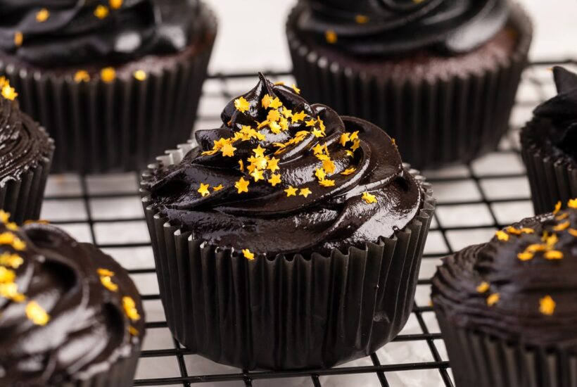 chocolate cupcake with black cream cheese frosting frosting and gold sprinkles on cooling rack.