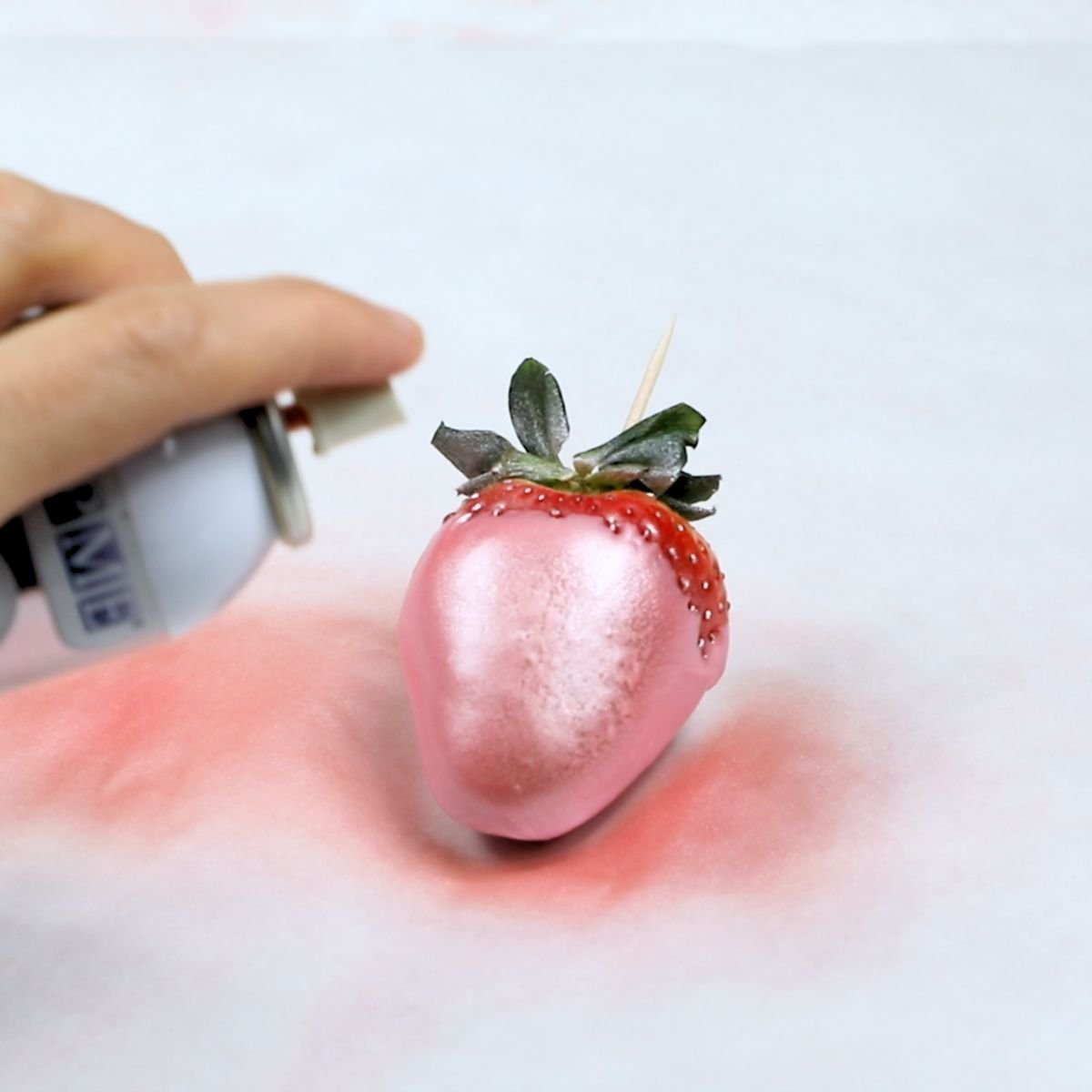 spraying pink edible luster dust onto a single pink chocolate dipped strawberry on parchment paper.