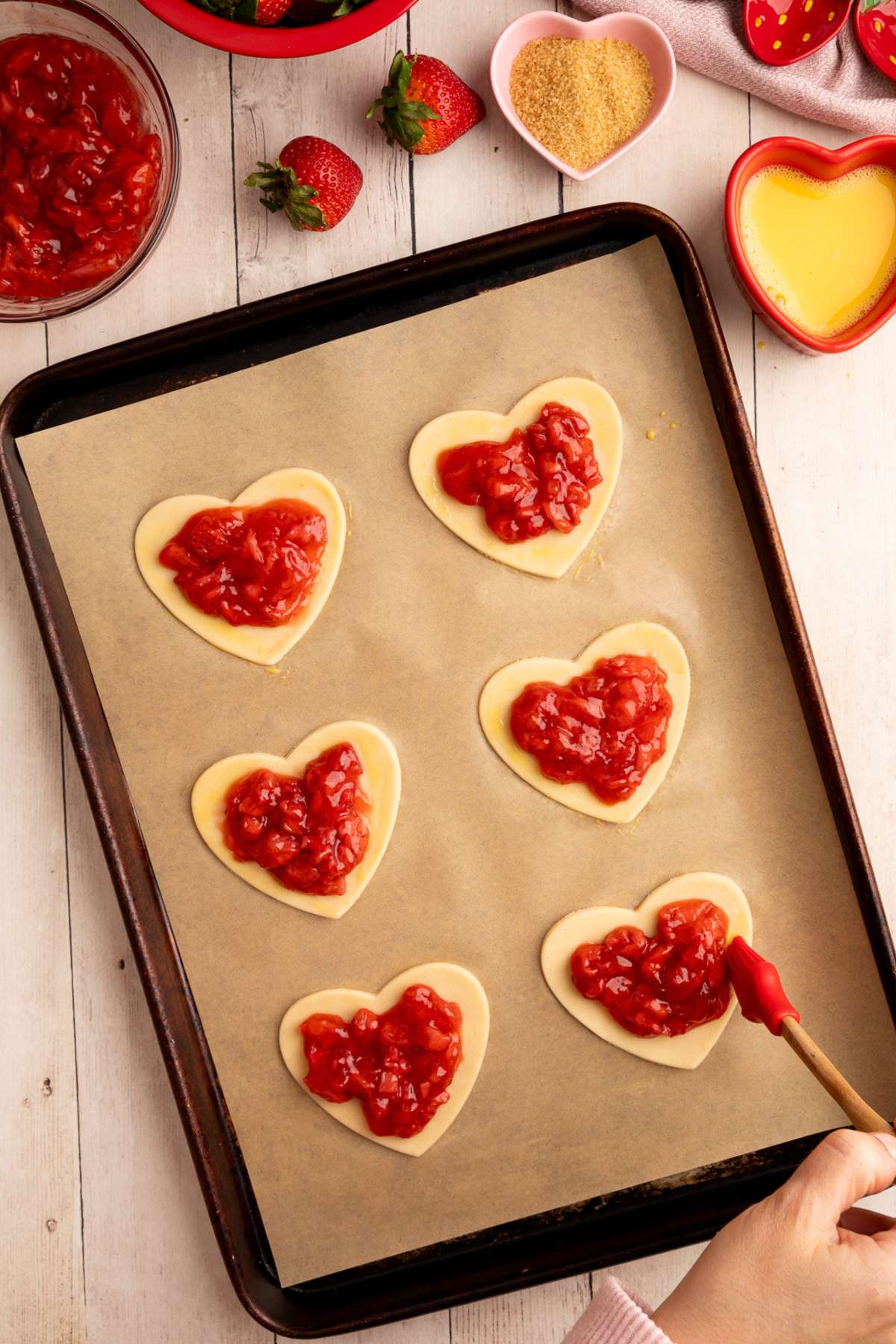 6 heart shaped hand pies on a baking sheet, with filling in the middle of the pies, and using a red pastry brush to brush on an egg wash around the edges of the crusts.