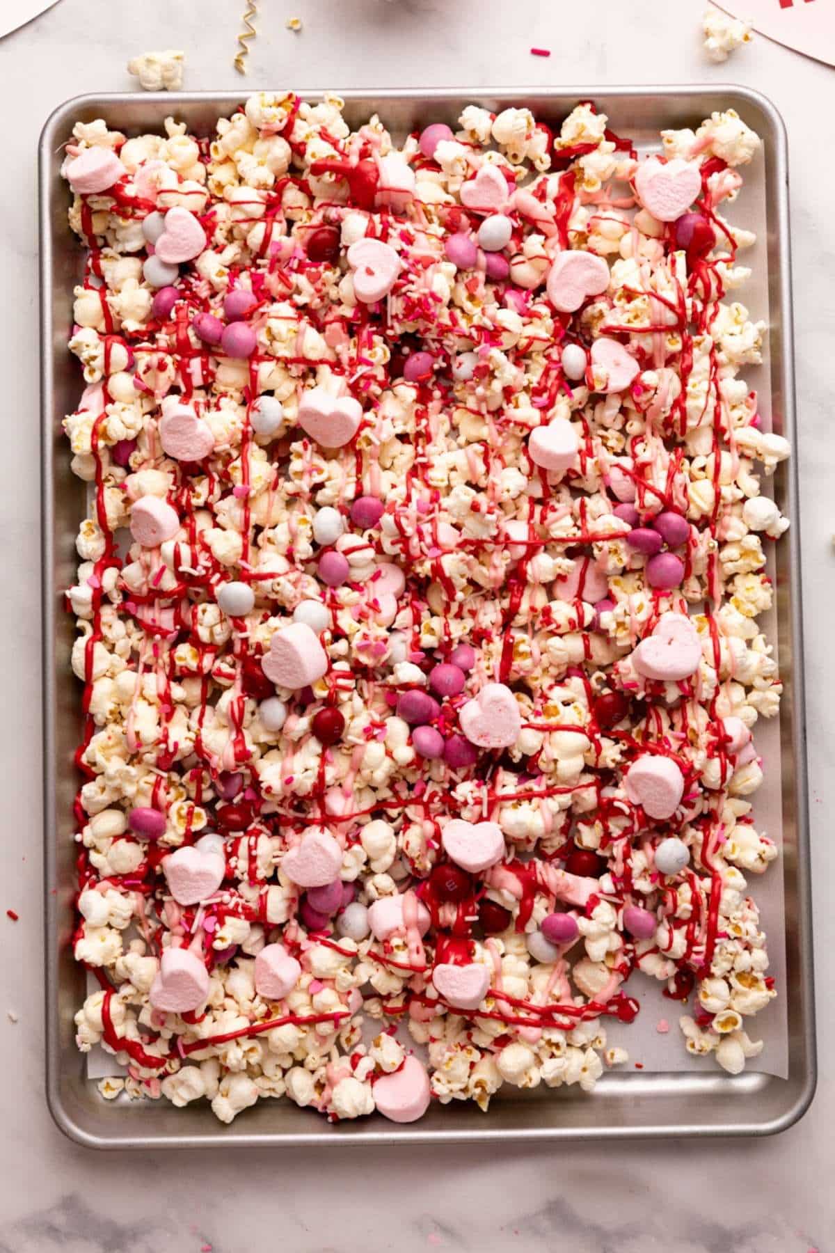 Adding more toppings and chocolate to Valentine's day popcorn on a baking sheet.