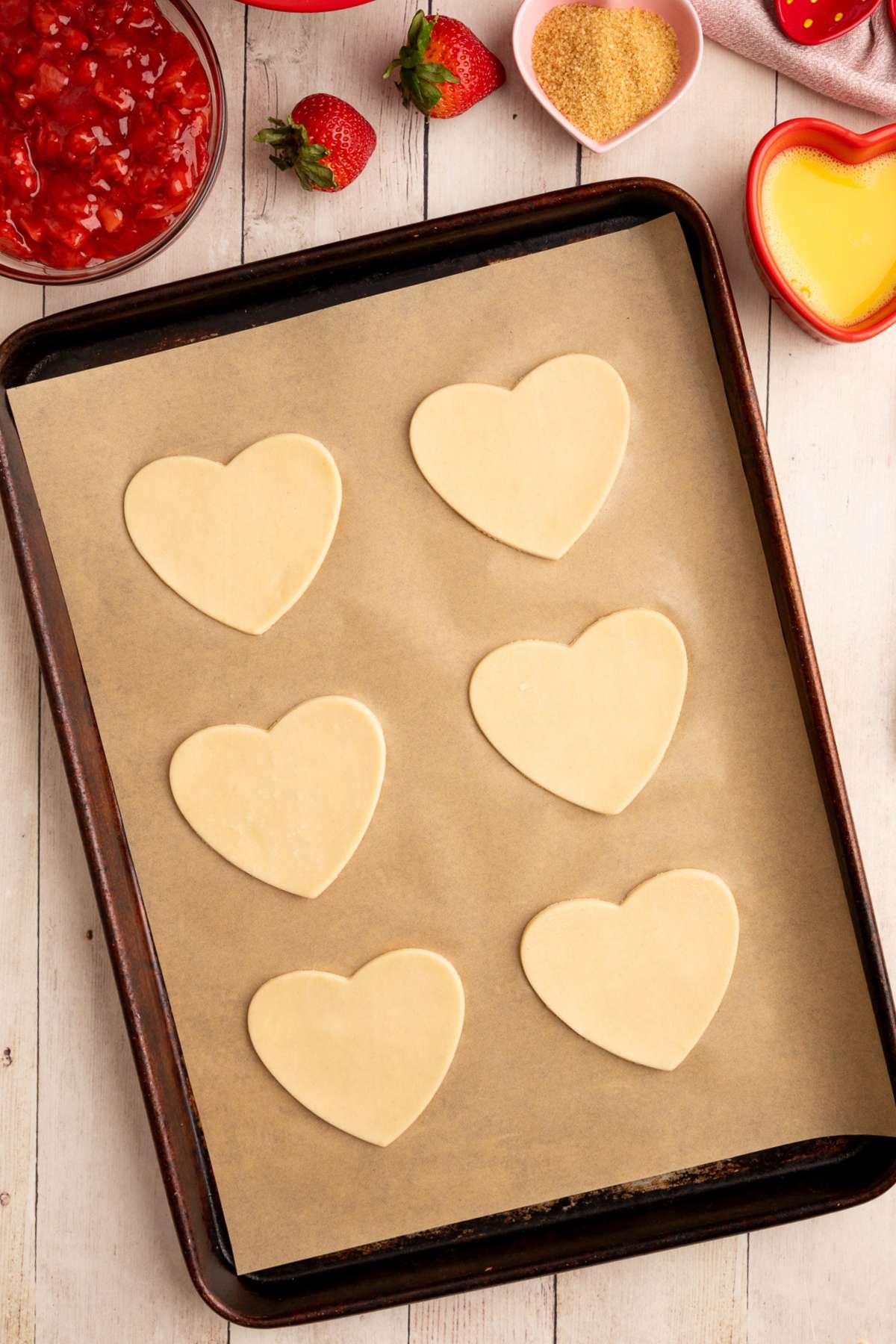 6 heart shaped unbaked pie crusts on a baking sheet lined with brown parchment paper.