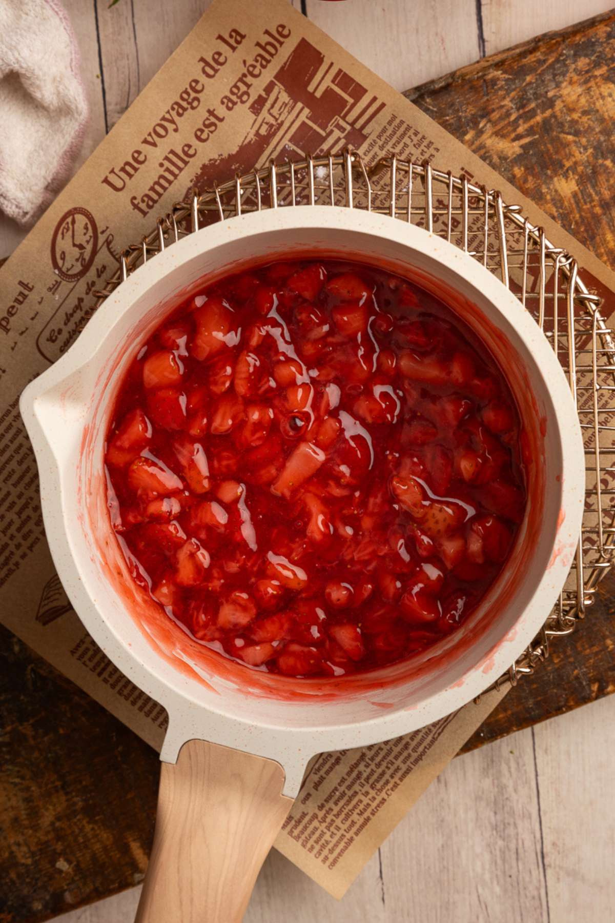 cooked strawberry pie filling in a cream saucepan with a wood handles on a cooling rack.