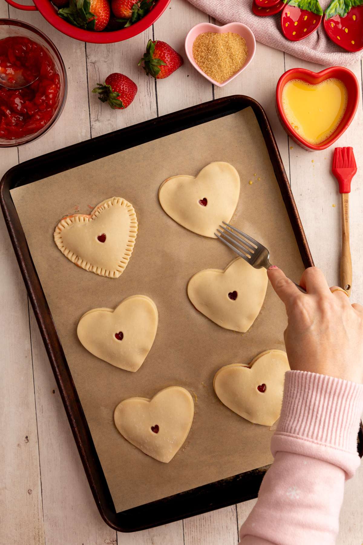 6 heart shaped hand pies on a parchment lined baking sheet, and using a fork to crimp the edges of one of the pies.