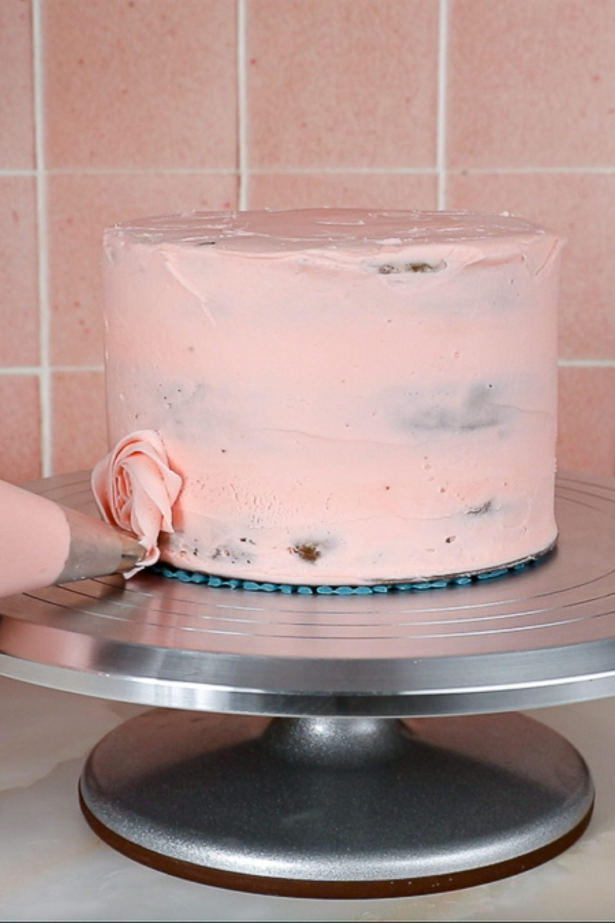 piping the first layer of pink rosettes onto the left side of a pink crumb coated cake on a metal cake turntable.