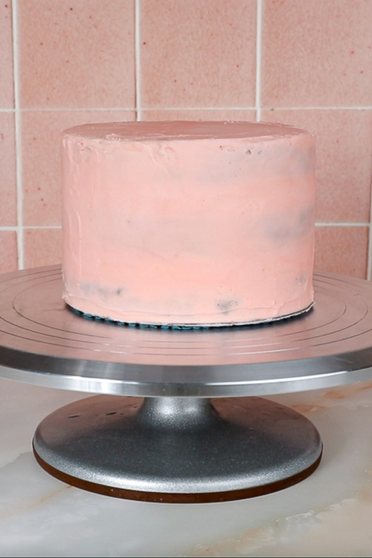 a three layer cake stacked, filled and crumb coated with pink frosting on a metal cake turntable.