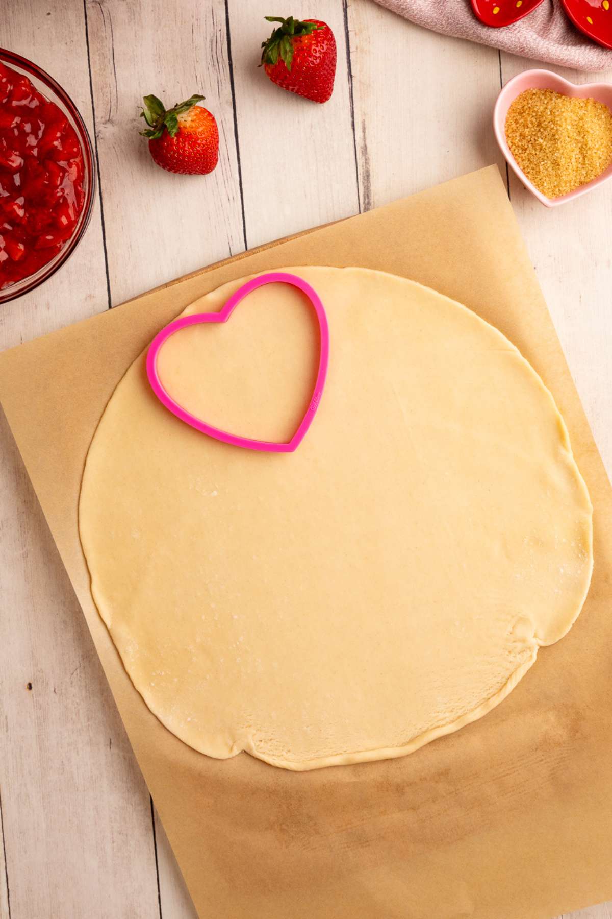 a pink heart shaped cookie cutter pressed into a rolled out round pie crust, on brown parchment paper.