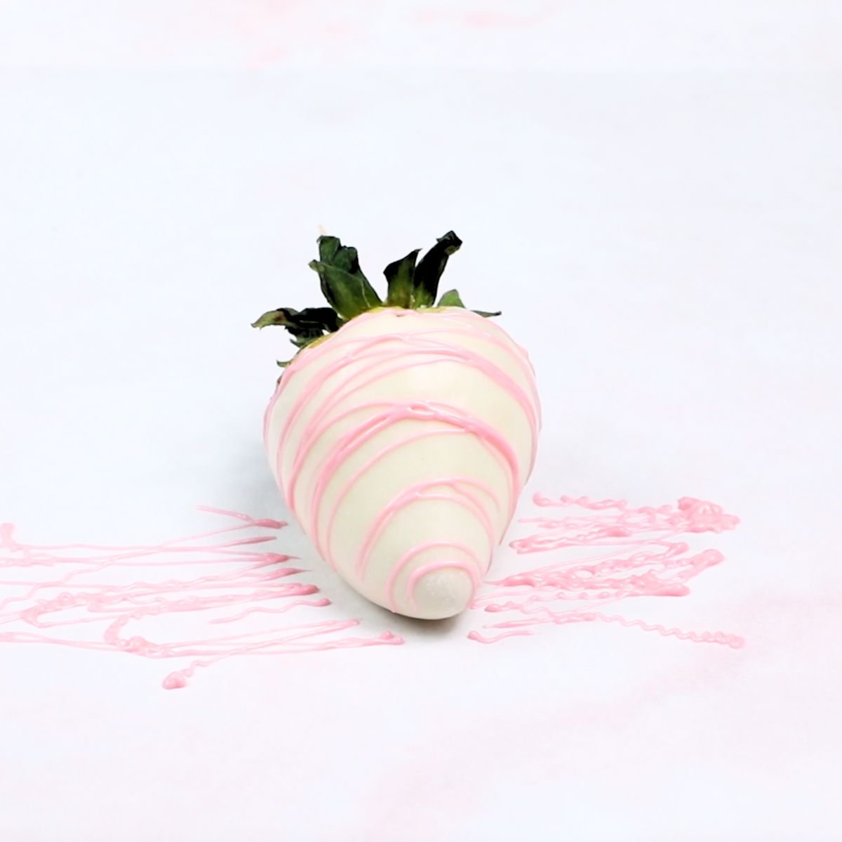 drizzling pink chocolate on a white dipped strawberry on a piece of white parchment paper.