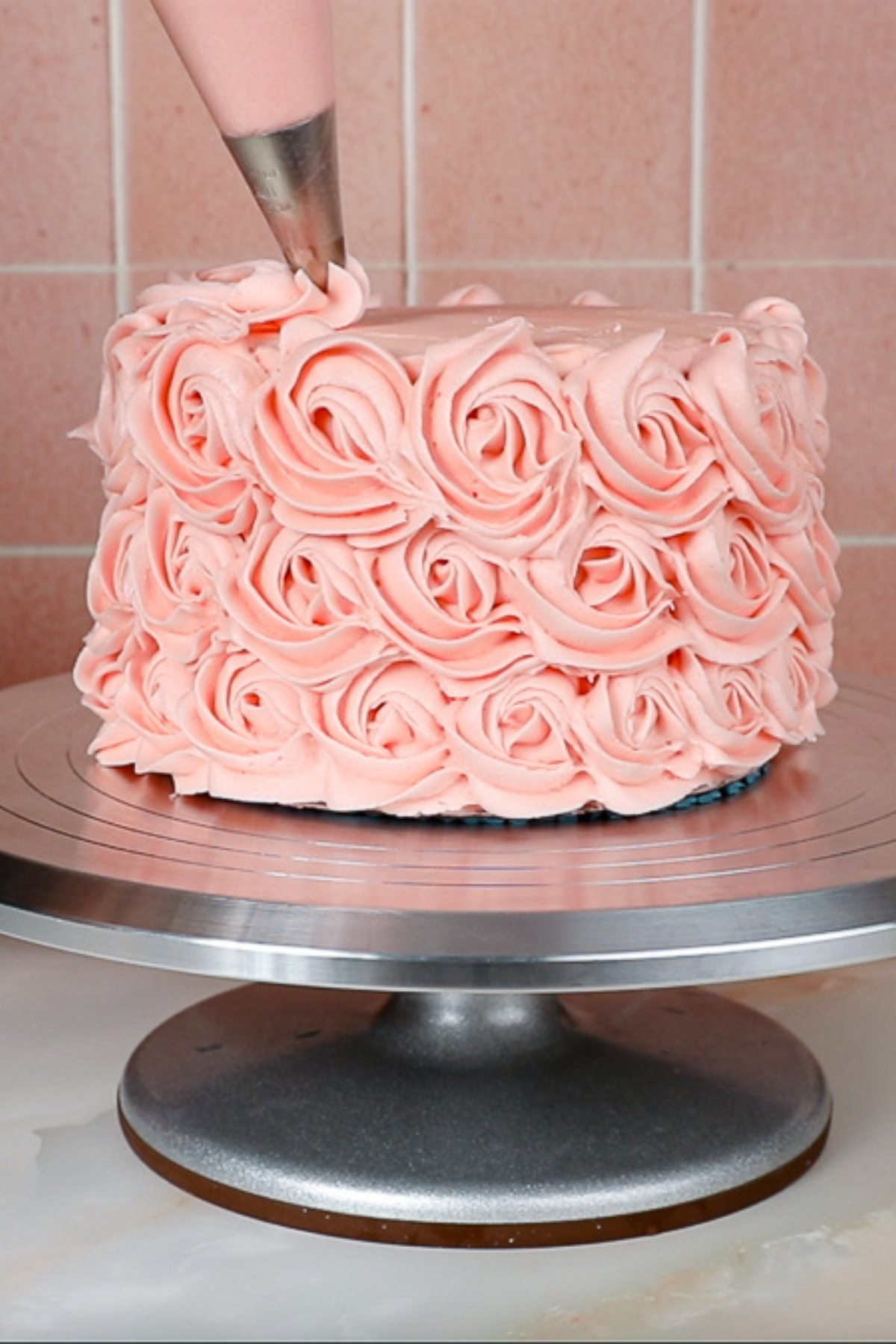 piping rosettes on to the top of a pink cake on a metal caketurntable.