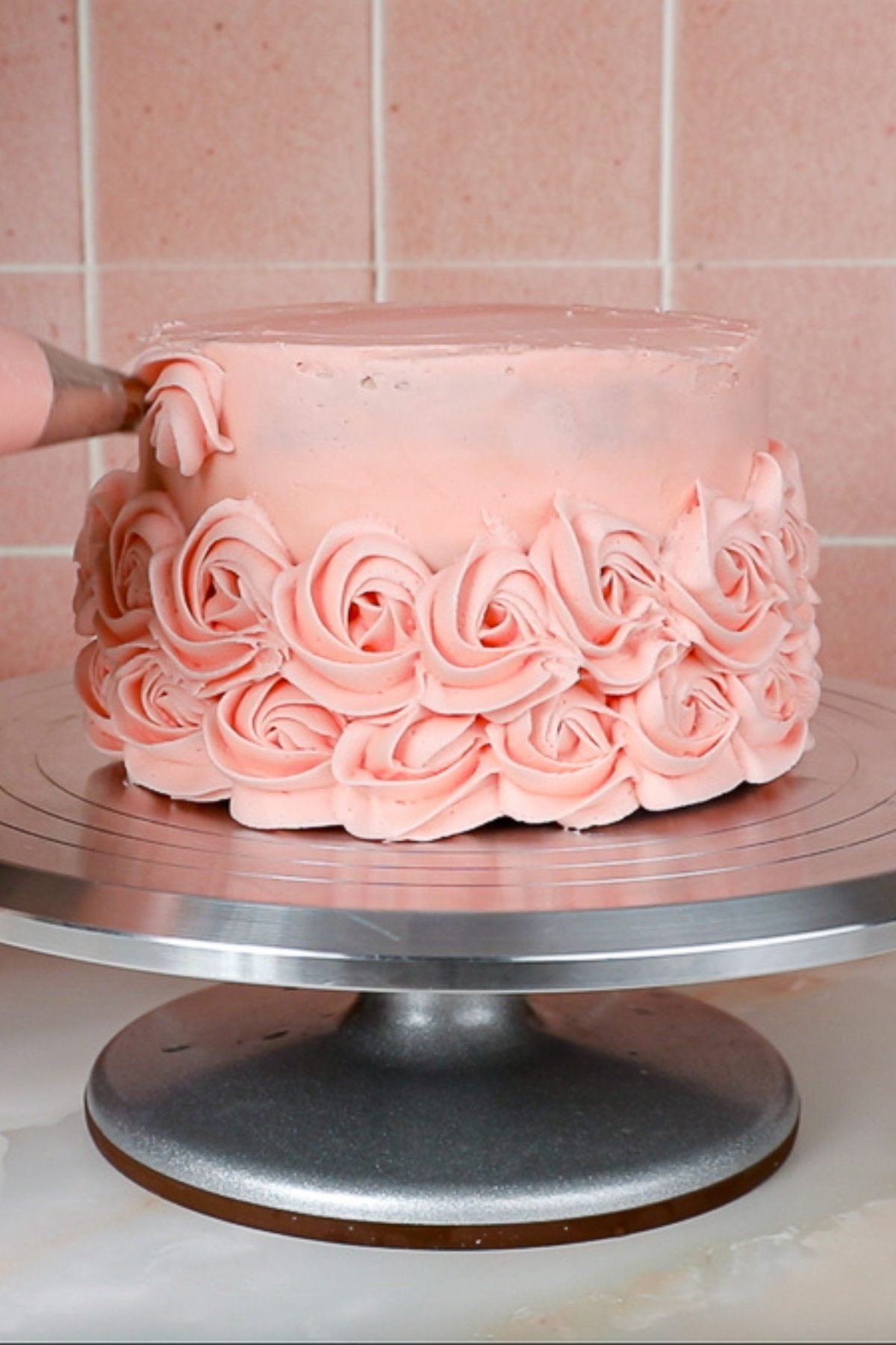 piping the third layer of pink rosettes onto a pink cake on a metal cake turntable.