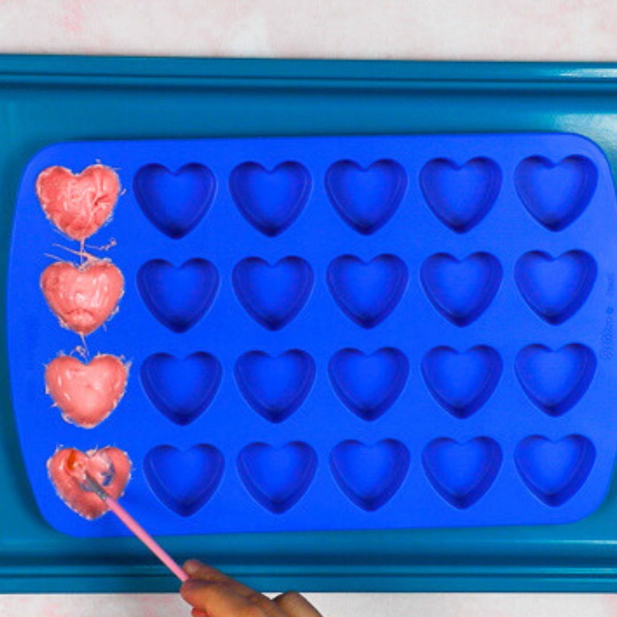 painting pink chocolate into the first row of a blue heart shaped silicone mold.