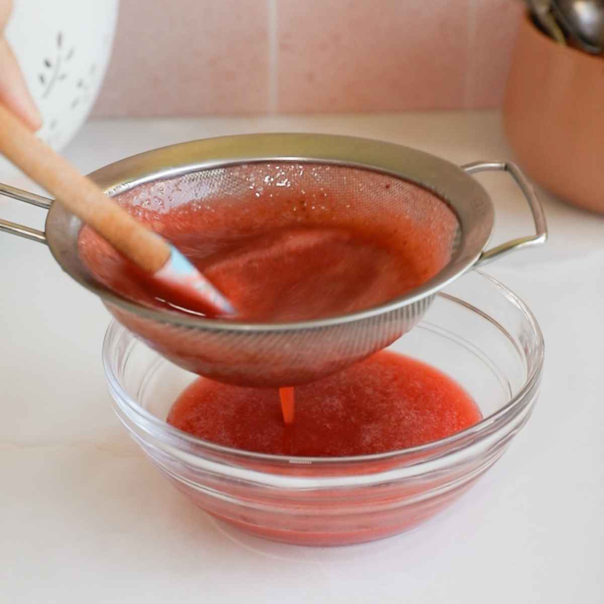 strainging strawberry syrup with a metal strainer over a glass bowl.