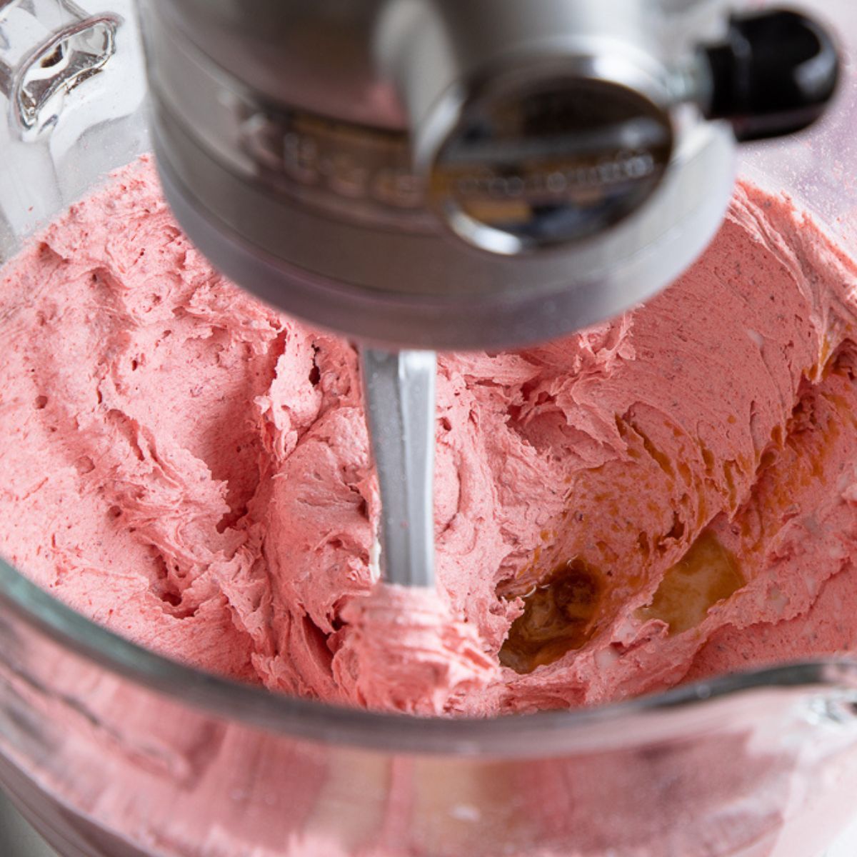 vanilla poured into a bowl of mixed strawberry frosting in a standing mixer.