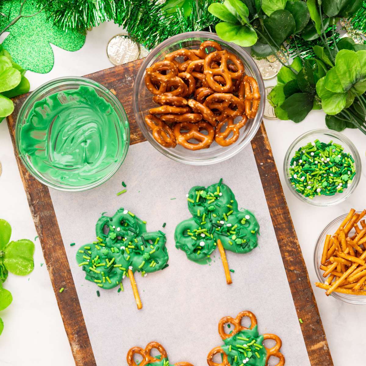 shamrock pretzels on a piece of parchment paper with sprinkles on top, along with bowls of pretzels, melted chocolate, and sprinkles.