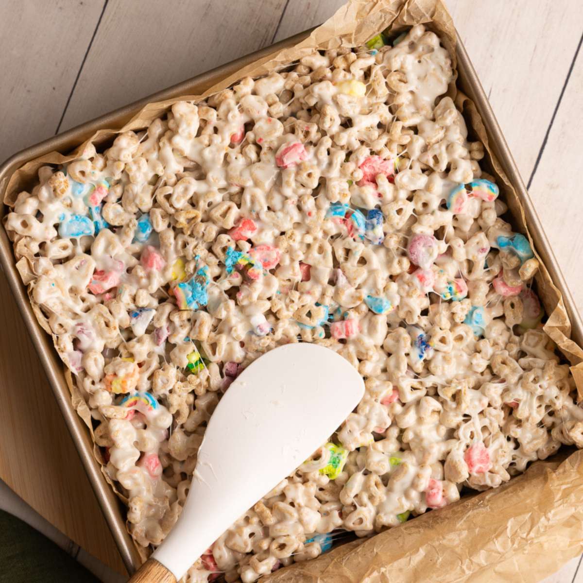 using a white spatula to press down lucky charms cereal bars into an 8x8 in pan lined with brown parchment.