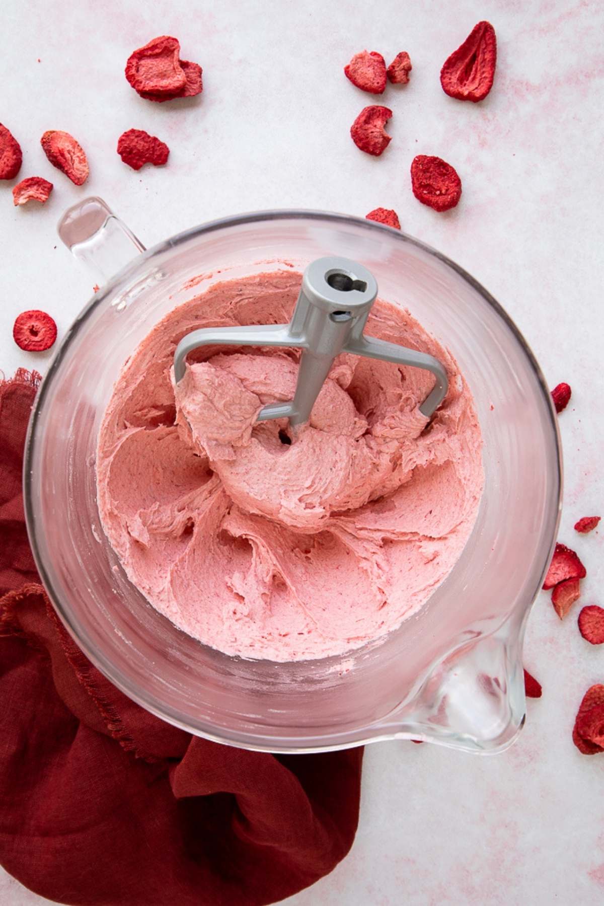 Strawberry Buttercream Frosting in a glass bowl with freeze dried strawberries and red tea towel around it.