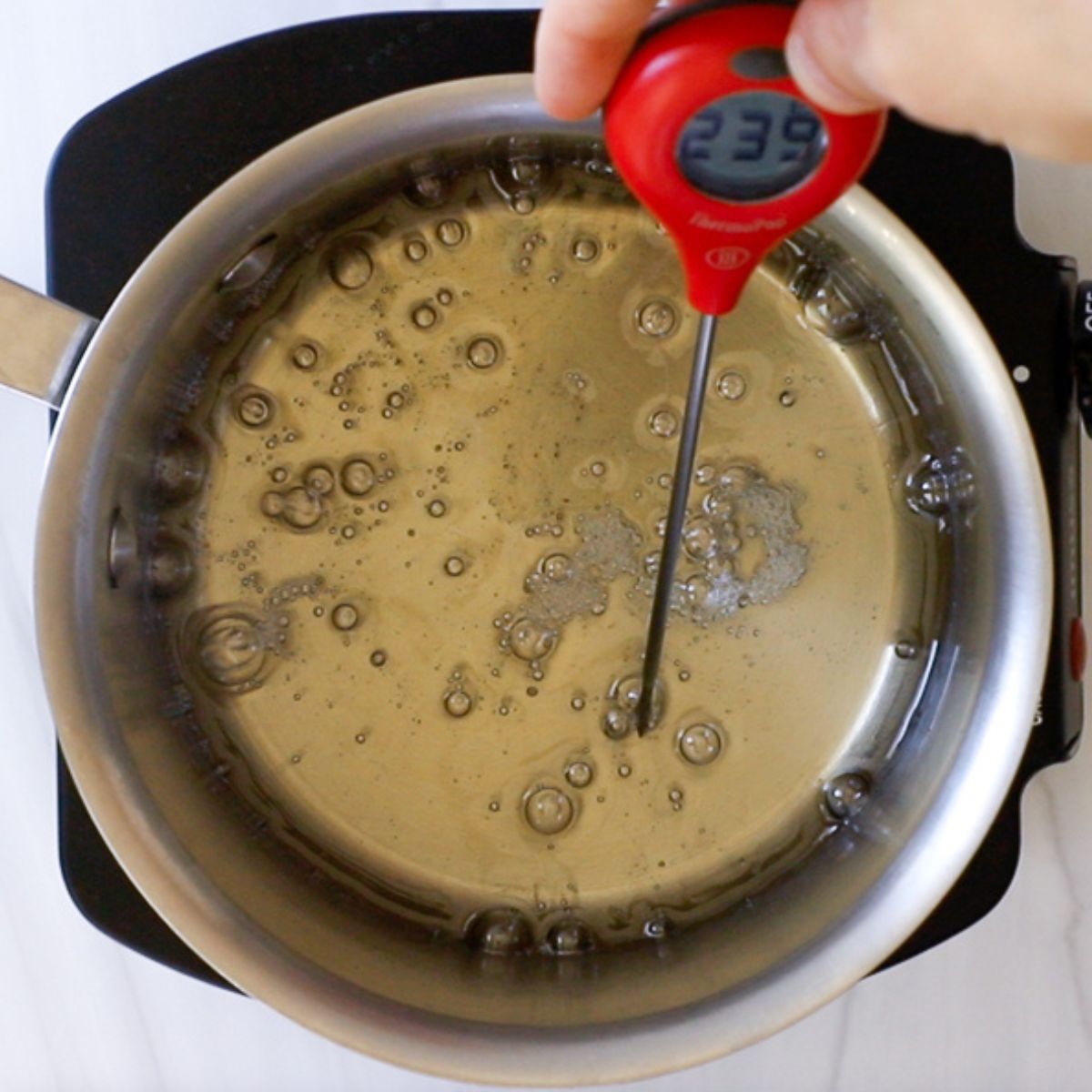 hand holding a red candy thermometer showing 239 ℉ in a saucepan of sugar syrup.