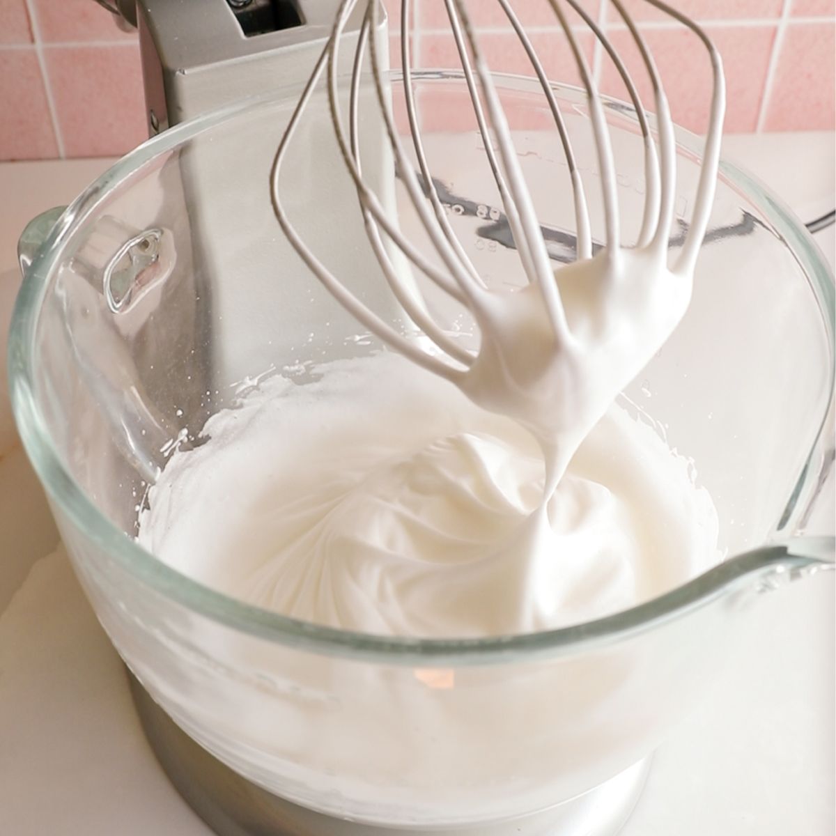 egg whites beat to soft peaks in the glass bowl of a grey standing mixer.